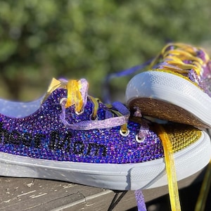 May include: Purple and gold sparkly sneakers with the words "Cheer Mom" on the side. The sneakers have yellow laces and a white sole.