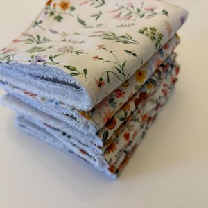May include: A stack of four square, floral-patterned cloths. The top cloth features a white background with delicate flowers in orange, purple, and green. The edges are white and fluffy. The other cloths have similar floral patterns.
