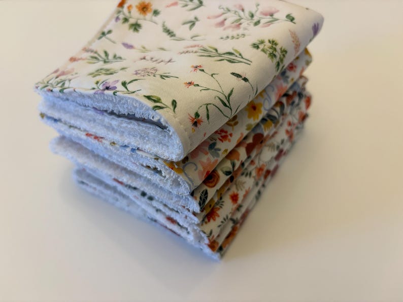 May include: A stack of four square, floral-patterned cloths. The top cloth features a white background with delicate flowers in orange, purple, and green. The edges are white and fluffy. The other cloths have similar floral patterns.