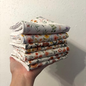 Può includere: Una pila di sei panni bianchi con stampa floreale e bordatura bianca. I panni presentano vari motivi floreali nei toni dell'arancione, giallo, rosa e verde. I panni sono tenuti in mano contro un muro bianco.