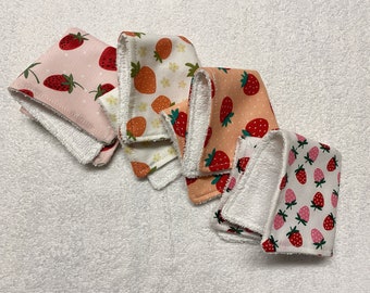May include: Five white washcloths with different strawberry patterns. The washcloths are folded and arranged in a row on a white fluffy surface.