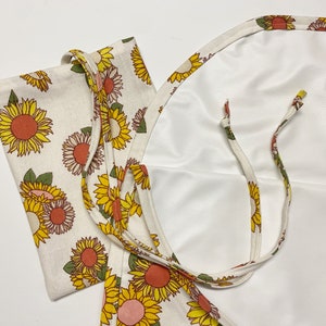 May include: A white fabric tote bag with sunflower print and two long straps. The bag is folded in half and the straps are tied together.