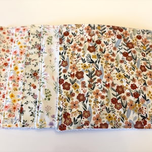 May include: A stack of floral patterned fabric squares. The fabric features a variety of colorful flowers in shades of red, pink, yellow, and brown, set against a white background. The edges of the fabric have a fringed trim.