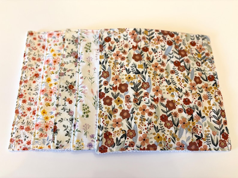 May include: A stack of floral patterned fabric squares. The fabric features a variety of colorful flowers in shades of red, pink, yellow, and brown, set against a white background. The edges of the fabric have a fringed trim.