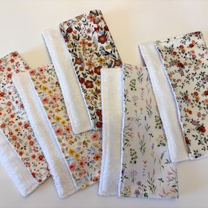 May include: A collection of six rectangular, floral-patterned cloths with white terrycloth borders. The fabric designs feature various flower patterns in shades of red, yellow, and purple on a white background. The cloths are arranged on a white surface.