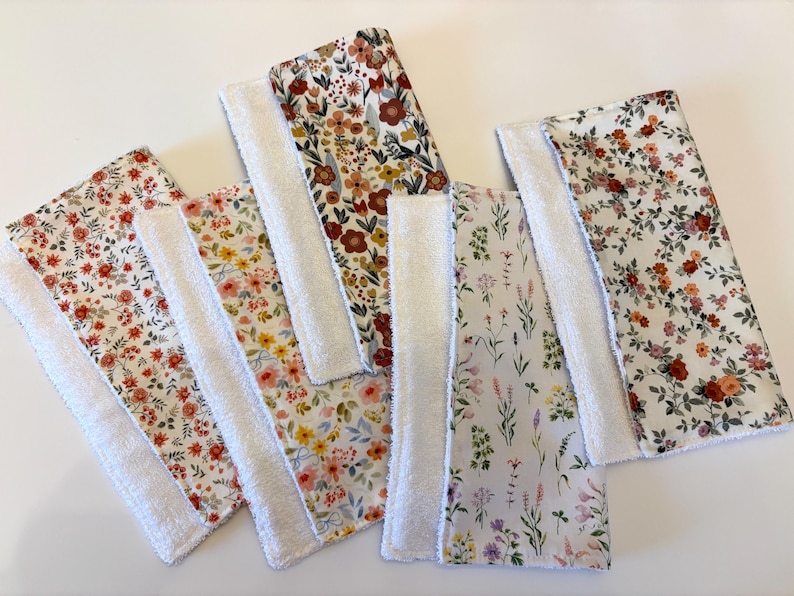 May include: A collection of six rectangular, floral-patterned cloths with white terrycloth borders. The fabric designs feature various flower patterns in shades of red, yellow, and purple on a white background. The cloths are arranged on a white surface.