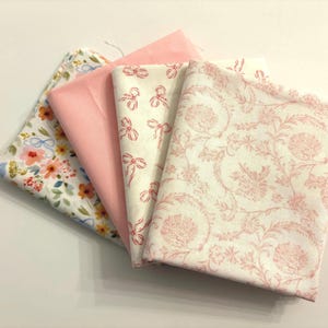 Puede incluir: Cuatro muestras de tela en tonos rosa y blanco. Una muestra presenta un estampado floral con lazos, otra es rosa liso y las otras dos tienen diseños de lazos y flores. Las telas están dobladas y superpuestas.