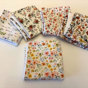 May include: A collection of six floral-patterned cloth napkins. The napkins feature various designs with flowers in shades of pink, orange, yellow, and brown, set against white backgrounds. Each napkin has a white, fringed edge.