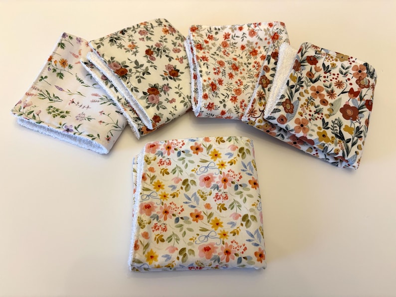 May include: A collection of six floral-patterned cloth napkins. The napkins feature various designs with flowers in shades of pink, orange, yellow, and brown, set against white backgrounds. Each napkin has a white, fringed edge.