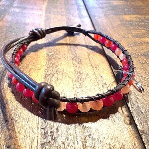 May include: A wrap bracelet featuring red and peach-colored beads, wrapped with dark brown leather and wire. The bracelet is knotted at the closure. The beads are a mix of opaque and translucent.
