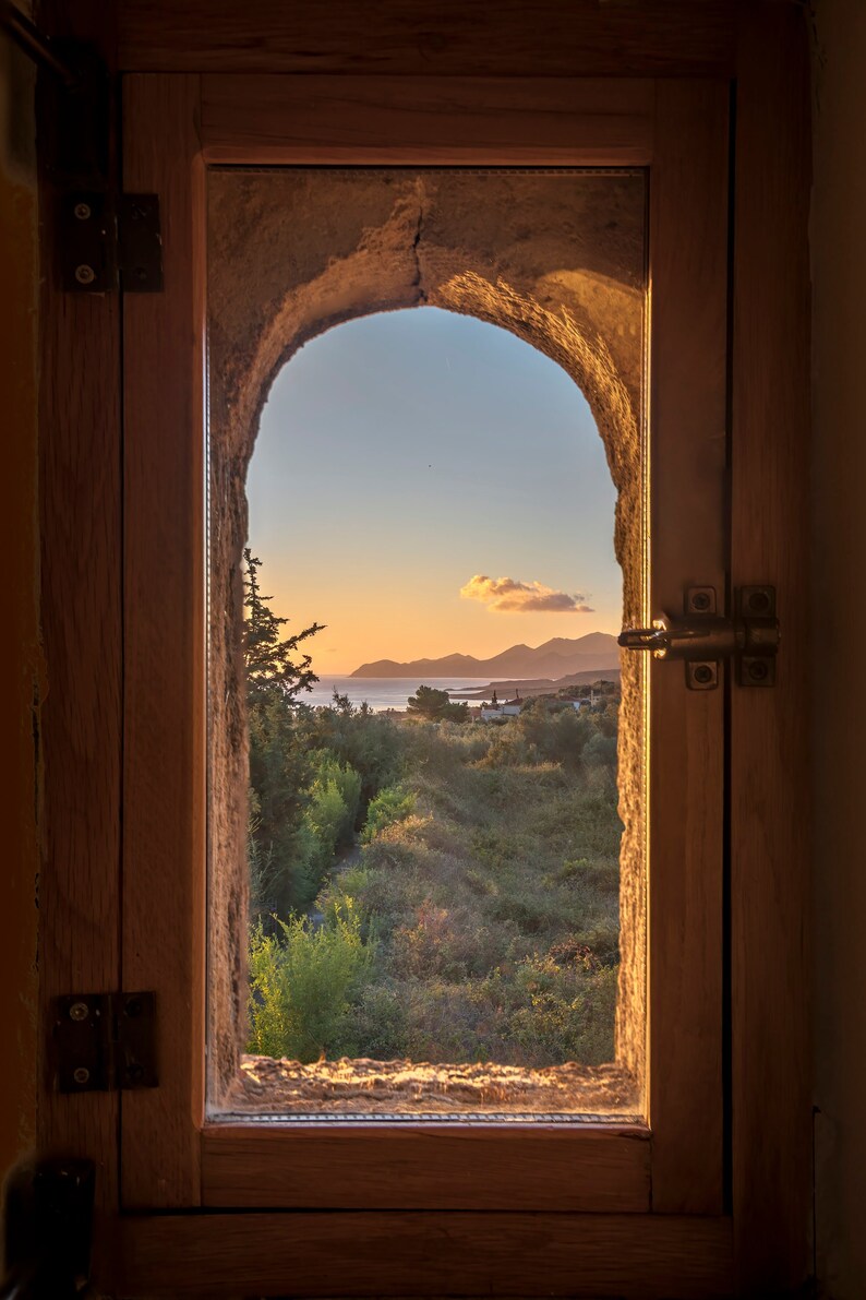 Puede incluir: Una ventana de madera con un marco de piedra arqueado ofrece una vista de una puesta de sol sobre un paisaje costero. La ventana tiene bisagras y un pestillo de metal. La vista incluye &aacute;rboles, colinas y una masa de agua.