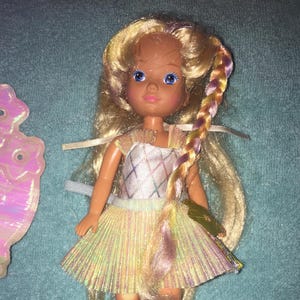 May include: A vintage doll with long blonde hair styled in a braid with pink and purple streaks. She is wearing a white and iridescent dress with a diamond pattern and a matching iridescent skirt.
