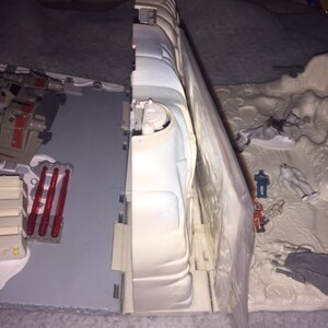 May include: A white plastic playset with a spaceship, a wall, and several figures. The spaceship is beige and gray with red accents. The wall is white and has a blue and white pattern. The figures are small and made of plastic. The playset is set on a gray surface.