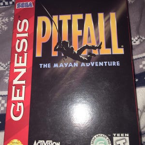 May include: A SEGA Genesis game box for Pitfall: The Mayan Adventure. The box is black with the game title in large yellow letters and a silhouette of a person swinging on a rope. The SEGA Genesis logo is on the left side.