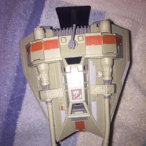 May include: A white and orange toy spaceship with a black stand. The spaceship has two large cannons on the bottom and a smaller cannon on the top.