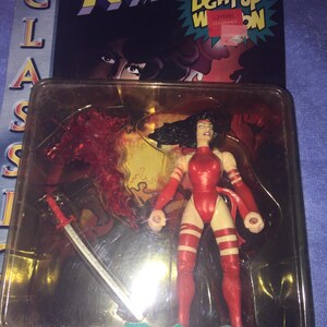 May include: A red and white action figure of Elektra from the X-Men comics, holding a light-up ninja blade. The figure is packaged in a plastic blister pack with a red and yellow X-Men logo and the words "Light Up Weapon" on the front.