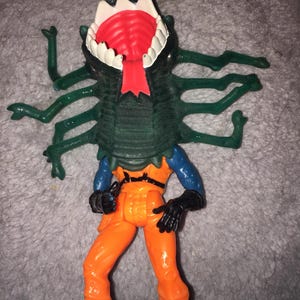 May include: A plastic action figure featuring a dark green, multi-legged creature head with a wide, open mouth, white teeth, and a red tongue, attached to a body wearing an orange jumpsuit, blue sleeves, and black gloves and boots.