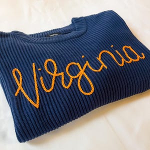 Puede incluir: Jersey de punto acanalado azul marino con la palabra "Virginia" bordada en hilo naranja. El jersey tiene cuello redondo y una etiqueta "Fashion". La prenda est&aacute; doblada sobre una superficie blanca.