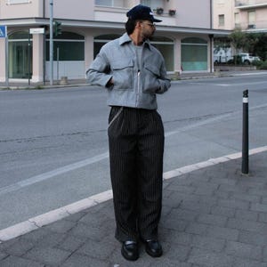 May include: A grey zip-up jacket with two front pockets and black and white striped trousers. The trousers have a silver chain attached to the belt loop. The person is wearing a navy blue baseball cap and black shoes.