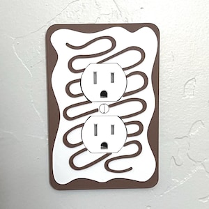 May include: A white electrical outlet cover with a brown border and a design of a chocolate-covered pastry with a swirl pattern.