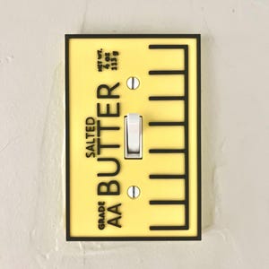 以下が含まれることがあります： 黒い縁取りのある黄色のライトスイッチカバー。カバーには「GRADE AA BUTTER SALTED」と「NET WT. 4 OZ. 113 g」の文字が印刷されています。