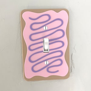 May include: A light switch cover with a pink background and purple frosting-like design. The switch is white and has a single toggle.