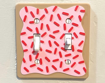 Puede incluir: Una tapa de interruptor de luz con un diseño de glaseado rosa y rojo. La tapa tiene dos interruptores de palanca.