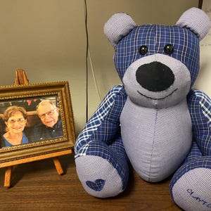 Puede incluir: Un oso de peluche confeccionado con varios parches de tela de cuadros y vichy en azul y blanco. El oso presenta ojos de botón negros, una nariz de tela negra, una sonrisa cosida y el nombre Clayton bordado en un pie con un corazón en el otro.