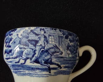 May include: A white and blue ceramic teacup with a handle. The cup features a blue transferware design depicting a person on horseback, trees, and a building. The cup's base is white, and the rim is blue.