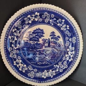 May include: A round ceramic plate with a cream-colored center and a wide, deep blue border. The center features a detailed landscape with trees, a stone bridge, and buildings. The border is decorated with white floral and geometric patterns.