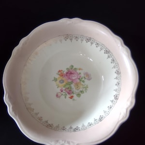 May include: A round ceramic bowl with a scalloped edge, featuring a pale pink rim and a white center. The interior is decorated with a central floral bouquet of pink, yellow, and orange flowers, surrounded by a delicate silver filigree border.