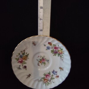 May include: A white porcelain saucer with a scalloped gold rim and colorful floral patterns. A measuring tape is positioned vertically against the saucer, showing a diameter of approximately 6 inches across the center of the dish.
