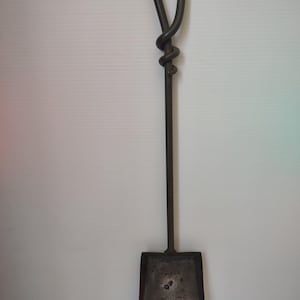 May include: A dark metal fireplace shovel with a square blade and a long handle. The handle features a decorative knot near the top and a loop at the end. The shovel is designed for removing ashes and embers.