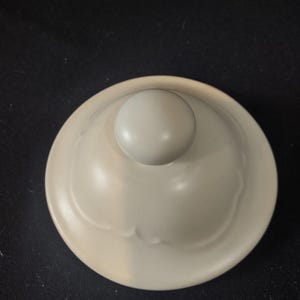 May include: A light beige ceramic lid with a round knob on top. The lid has a smooth, curved surface with subtle decorative indentations. The background is a dark, solid color.