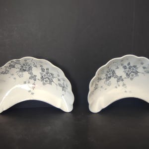 May include: Two white, scalloped-edge serving dishes with a floral pattern in shades of gray. The dishes have a curved, boat-like shape, and are set against a dark background. The floral design is delicate and covers the surface of the dishes.