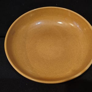 May include: A round, shallow bowl with a smooth, solid mustard-yellow finish. The interior surface features a subtle, speckled texture throughout, and the rim is slightly raised, creating a simple, circular profile for this ceramic dish.