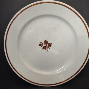 May include: White ceramic plate with a brown leaf design in the center. The plate has a brown rim and a brown border. This plate is suitable for serving food.