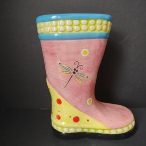 May include: A decorative ceramic boot vase, painted in pink, yellow, and blue. The boot features a dragonfly design and polka dots. The top rim is blue with yellow accents. The sole is black.