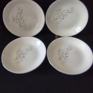 May include: A set of four round white ceramic bowls featuring a silver rim and a delicate floral pattern in shades of blue and gray. Each bowl has a smooth, glossy finish and a curved edge, displaying a consistent botanical design on the surface.