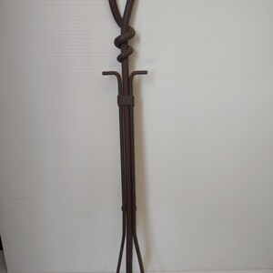 May include: A wrought iron fireplace poker with a looped handle and a decorative knot detail. The poker has a vertical shaft with a horizontal bar and three curved legs at the base. The metal has a dark brown finish.
