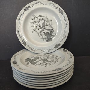 May include: A stack of white ceramic plates with a gray bird and floral design. The plates have a decorative border with small floral accents. The plates are stacked on a dark surface.