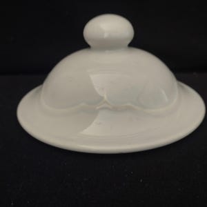 May include: A white ceramic lid with a rounded top and a small knob. The lid has a scalloped edge and a glossy finish, reflecting the surrounding environment. The object is likely a replacement lid for a teapot or serving dish.