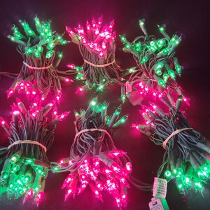 May include: Six bundles of string lights with dark green wire and small incandescent bulbs in alternating bright pink and vibrant green colors. Each bundle is secured with a tan rubber band, and one white electrical plug is visible at the bottom.