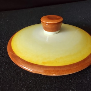 May include: A round ceramic lid featuring a glossy finish with a gradient color scheme. The center is a pale cream color that transitions into a vibrant yellow band, finished with a dark orange-brown rim and a matching cylindrical knob handle.