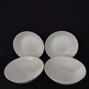 May include: A set of four plain white ceramic bowls arranged in a two-by-two grid. Each bowl features a smooth, rounded interior and a simple, clean design with a consistent glossy finish across all four pieces, presented against a dark background.