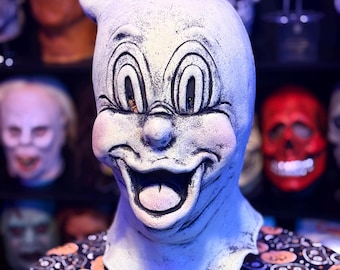 May include: A white, smiling mask with large eyes and a prominent nose, reminiscent of a cartoon character. The mask is set against a backdrop of other masks, suggesting a Halloween or costume theme. The person is wearing a black shirt with a Halloween pattern.