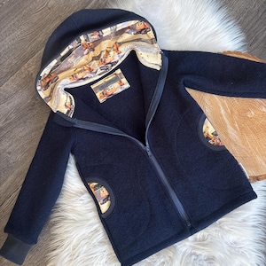 Könnte beinhalten: Marineblaue Kapuzenjacke mit durchgehendem Reißverschluss. Das Innenfutter und die Details der Taschen zeigen einen farbenfrohen Druck von Zeichentrickfahrzeugen. Die Jacke wird auf einer weißen Kunstfelloberfläche präsentiert.