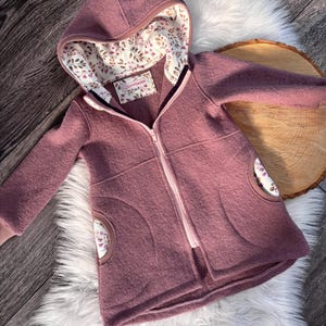 Könnte beinhalten: Staubrosa Kapuzenjacke mit durchgehendem Reißverschluss. Die Jacke hat ein floral gemustertes Futter in der Kapuze und in den Taschendetails. Sie wird auf einem weißen Kunstfellteppich und einer Holzoberfläche präsentiert.