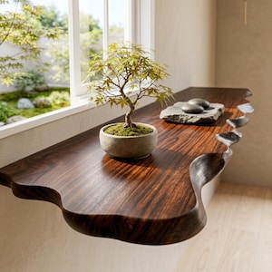May include: A dark brown wooden wall shelf with a natural live edge featuring a wavy, carved front profile. A small green bonsai tree in a round, light-coloured ceramic pot and two smooth grey stones on a flat rock sit on the shelf surface.