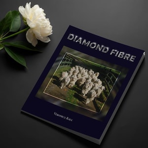 May include: A book titled "DIAMOND FIBRE" with a dark blue cover, featuring a photograph of sheep. A white peony flower and green leaves are in the upper left corner. The author's name, Veronica Rall, is at the bottom.